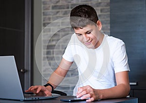 Cute young teenager in white shirt sitting behind desk in kitchen next to laptop and study.