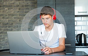 Cute young teenager in white shirt sitting behind desk in kitchen next to laptop and study. Serious boy in earphones