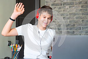 Cute young teenager in white shirt sitting behind desk in kitchen next to laptop and study. Serious boy in earphones