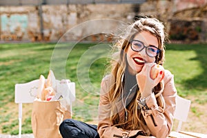 Cute young white woman with red apple in hand dreamily looks at camera while sitting in the park after shopping. Pretty