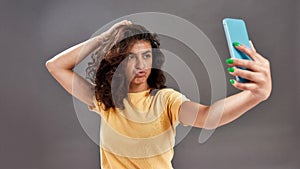 Cute young woman with curly dark hair making selfie photo with smartphone isolated over gray background