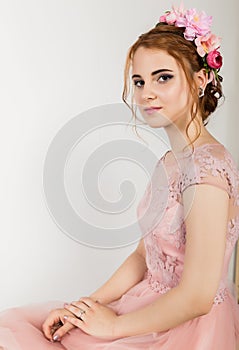 Cute young woman in delicate pink dress sits on sofa, professional hairstyle with wreath.