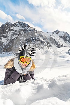 Cute young woman dressed in winter clothes portrait, glasses and winter funny hat, resting on snow top of the mountain