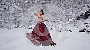 Cute young woman in fairy tale image in red royal dress with white violin stands on snow in winter forest