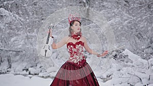 Cute young woman in fairy tale image in red royal dress with white violin stands on snow in winter forest