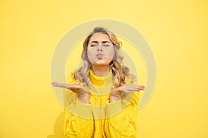 Cute young woman in knitted sweater blowing a kiss from hand on yellow background, studio shot, thank you, merci