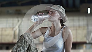 Cute young woman in military uniform drinking water from the bottle sitting on the floor in dusty dirty abandoned