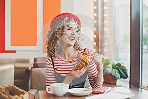 Cute young woman in red beret having a french breakfast with coffee and croissant, sitting at the cafe and laughing