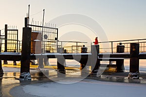 Cute young woman in red warm jacket looks at beautiful sunrise standing on snow-covered metal river pier