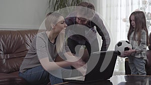 Cute young woman sitting on the leather sofa and bearded man standing near her working with laptop. Little girl comes