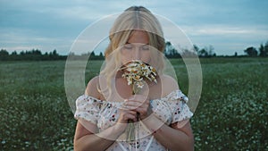 A cute young woman in a summer dress stands in a field and holds a bouquet of daisies