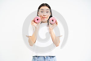Cute young woman with two tasty doughnuts, tempted to try glazed donnuts, white background