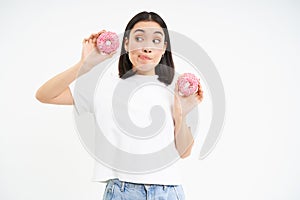 Cute young woman with two tasty doughnuts, tempted to try glazed donnuts, white background