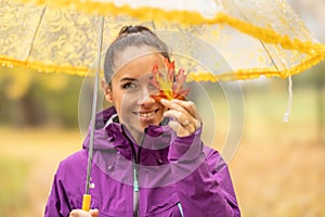 Cute young woman under an umbrella holding a colorful maple leaf and covering her eye in an autumn park