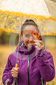 Cute young woman under an umbrella holding a colorful maple leaf and covering her eye in an autumn park