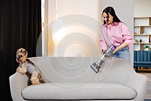 A cute young woman is vacuuming a couch with her cute dog sitting on it. Concept cleaning at home