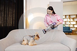 A cute young woman is vacuuming a couch with her cute dog sitting on it. Concept cleaning at home