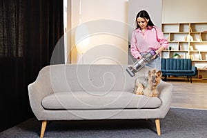 A cute young woman is vacuuming a couch with her cute dog sitting on it with a cordless vacuum cleaner at home