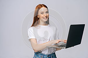 Cute young woman wearing T-shirt and denim pants holding laptop computer and looking at camera