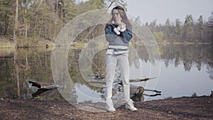 Cute young woman in white pants, jeans jacket and sunglasses standing on the riverbank. The girl is cold, she rubbing