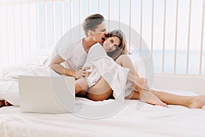 Cute young woman in white shirt sitting on bed and smiling, while boyfriend playfully bites her ear. Portrait of