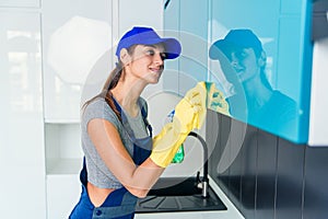 Cute young woman in workwear and protective rubber yellow gloves cleaning the furniture in modern hi-tech kitchen.