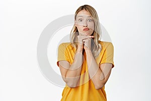 Cute young woman yearning, looking with interest, wishing smth, standing in yellow t-shirt over white background