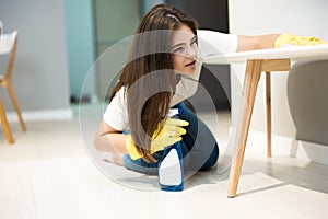 Cute young woman in yellow gloves with detergent spray in her hand wiping dust off from the kitchen chair with a rag