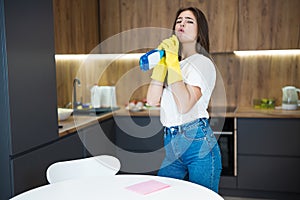 Cute young woman in yellow gloves holds jokingly spray detergent as a gun while wiping dust off from the kitchen table