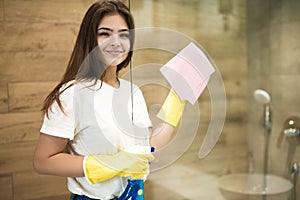 Cute young woman in yellow gloves spraying detergent spray while wiping dust off from the shower door with a rag in the