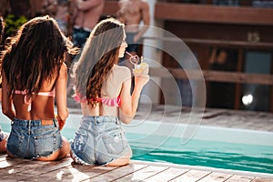 Cute young women sitting near swimming pool and drinking juice