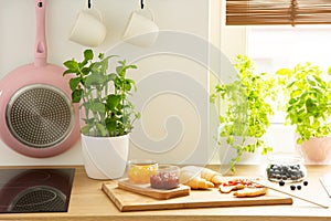 Cutting board with French croissants and fruit jams and natural herbs in a bright kitchen interior with a white wall