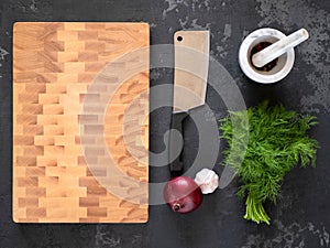 Cutting board, knife, onion, garlic, dill and mortar with spices on dark background, flatlay. Wooden block chopping board