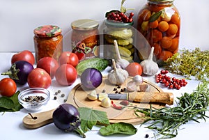 Cutting board with slices of garlic hot pepper, knife and eggplant, tomatoes, seasonings, herbs and jars of vegetables in the