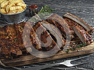 Cutting grilled pork ribs with sauce on a board, french fries, spices, wooden background