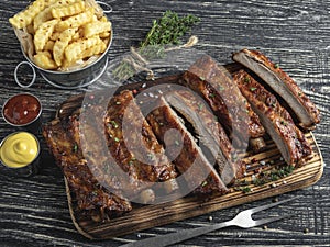 Cutting grilled pork ribs with sauce on a board, french fries, spices, wooden background