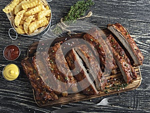Cutting grilled pork ribs with sauce on a board, french fries, spices, wooden background