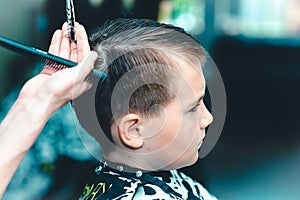 Cutting hair for young boy in a beauty salon.Hairdresser`s hands holding a comb and a colorful scissors.Closeup.Toned
