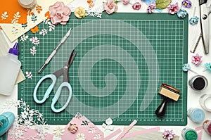 The cutting mat is surrounded by paper flowers, paper, tools and scrapbooking materials