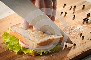 Cutting sandwich with bread, cheese, salad and ham with hands on wooden cutting board with knife