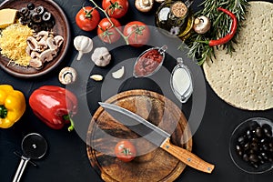Cutting wooden board with traditional pizza preparation ingredients: cheese, tomatoes, sauce, olives, olive oil, pepper