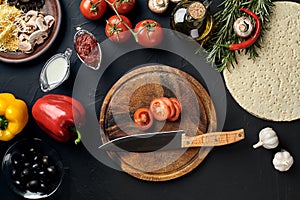 Cutting wooden board with traditional pizza preparation ingredients: cheese, tomatoes, sauce, olives, olive oil, pepper