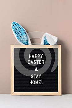 Cyan bunny ears and black felt letter board with a slogan - Happy Easter and Stay at Home on a beige background