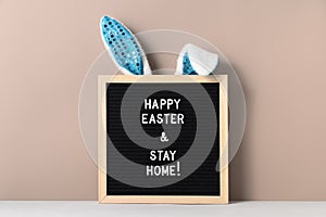 Cyan bunny ears and black felt letter board with a slogan - Happy Easter and Stay at Home on a beige background