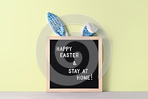 Cyan bunny ears and black felt letter board with a slogan - Happy Easter and Stay at Home on pastel green background