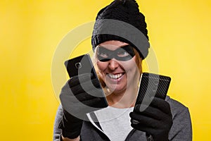 Cybercrime and money theft. A woman in a hat and mask with a malicious smile, holding phones. Yellow background. Copy space