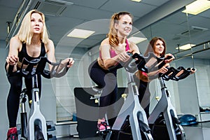 Cycling on exercise bikes. Three attractive young women in sports clothing exercising on gym bicycles.