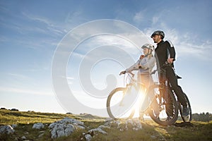 Cyclist couple with mountain bikes standing on the hill under the evening sky and enjoying bright sun at the sunset.