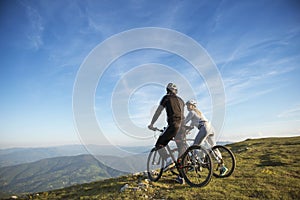 Cyclist couple with mountain bikes standing on the hill under the evening sky and enjoying bright sun at the sunset.