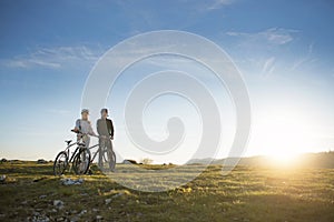 Cyclist couple with mountain bikes standing on the hill under the evening sky and enjoying bright sun at the sunset.